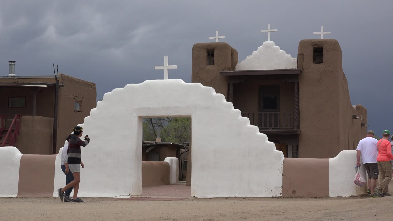Two friends pass in front of the central opening in the white church wall in Taos New Mexico, with brown buildings and crosses visible behind them.