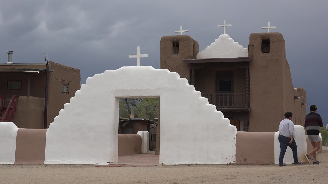 White adobe chapel gateway with two people near the entrance under cloudy sky.