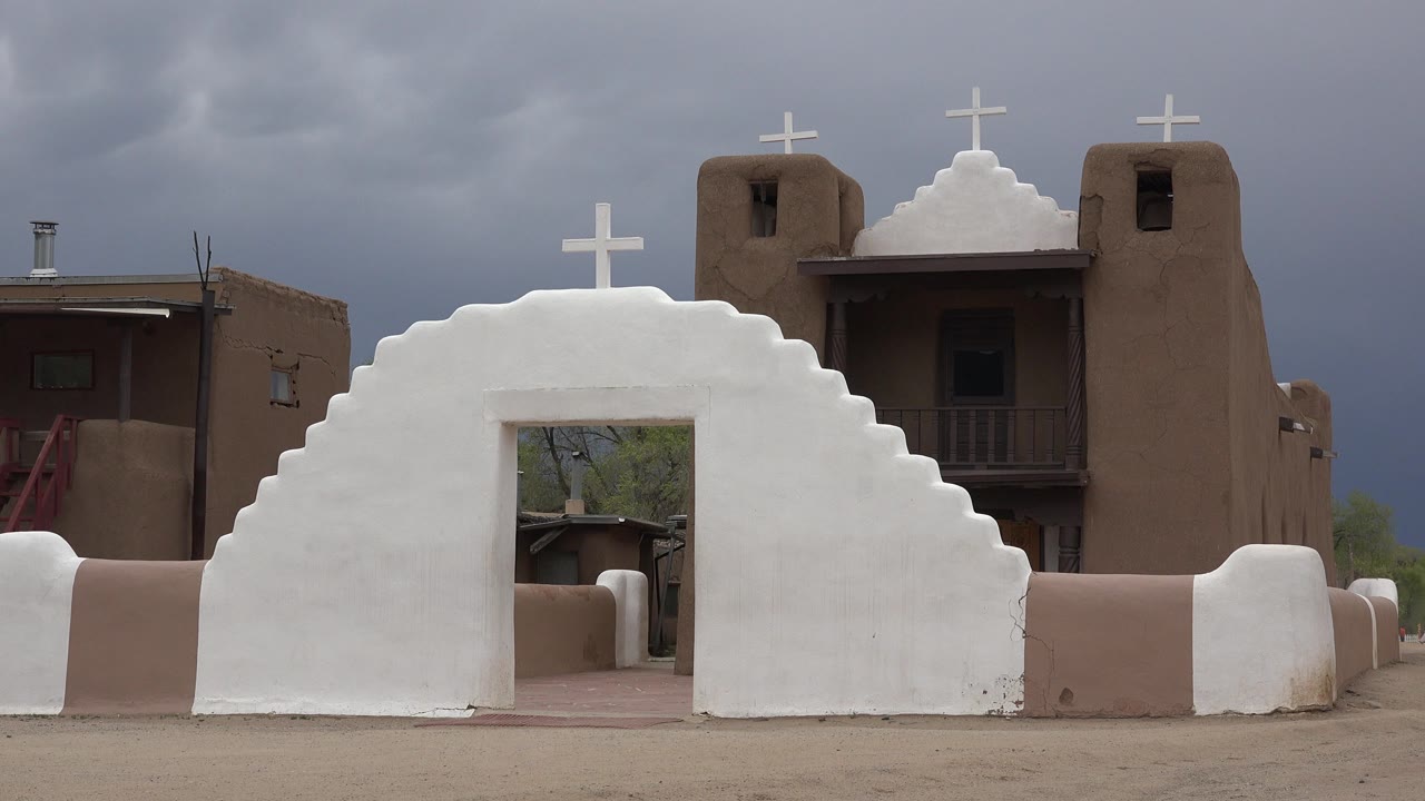 White stepped church wall with cross in front of brown adobe-style buildings.