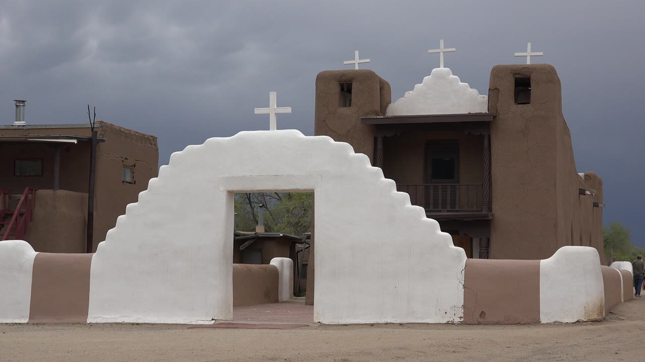Opening frame of a white stepped adobe church entrance in Taos New Mexico, with tan adobe towers, crosses, dark windows, gray clouds, and no visible people.