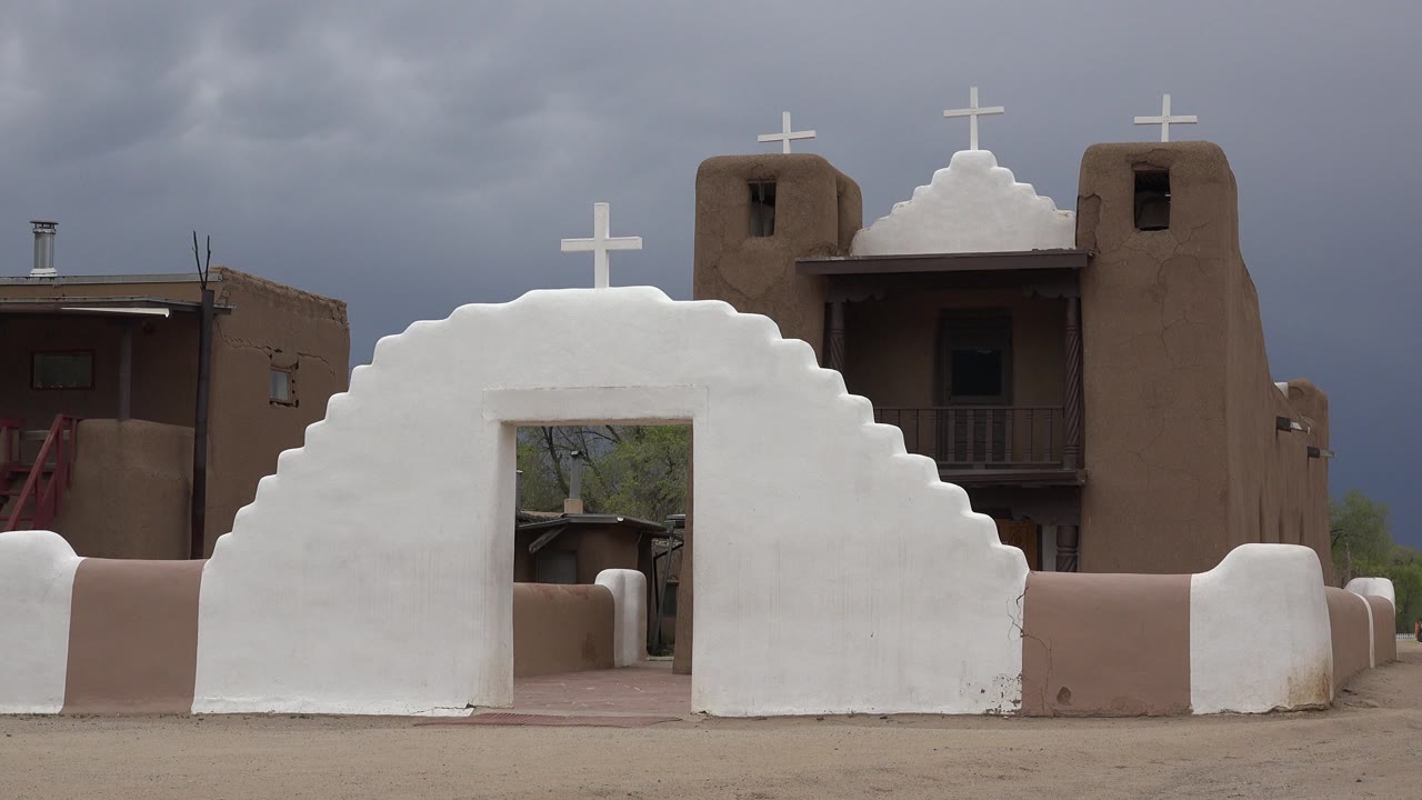 Opening frame of a white stepped wall with a cross in front of a brown religious building in Taos New Mexico, with dark clouds above and no visible people.