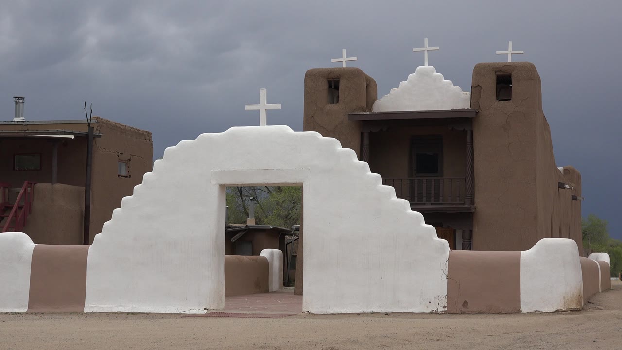 White adobe-style arch and tan religious building in Taos New Mexico appear under dark gray clouds at the start of the clip, with no visible people.