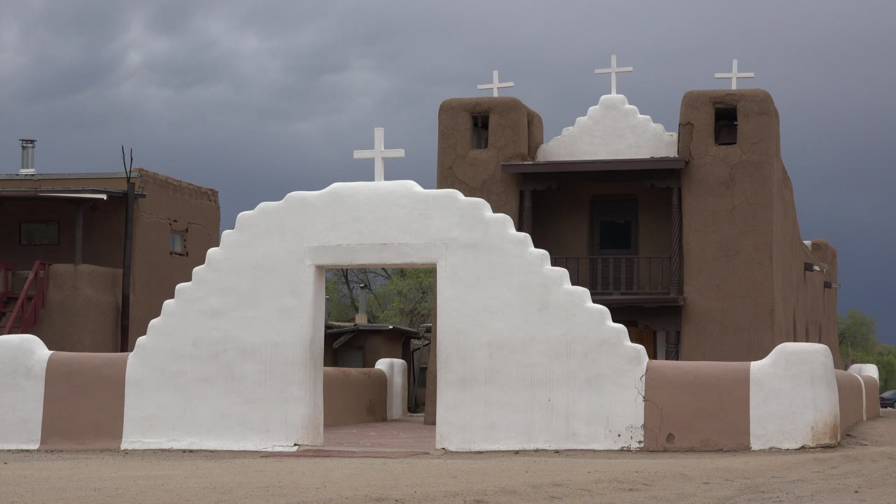 White stepped church wall with crosses and brown adobe-style buildings under gray clouds.
