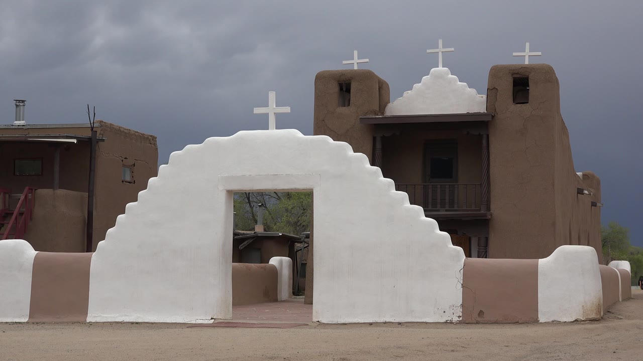 White adobe church wall with crosses and tan buildings under gray clouds.