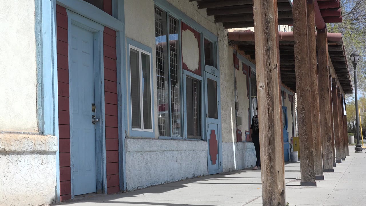Taos, New Mexico covered storefront walkway at the start of the clip, with a blue door in the foreground, wood posts along the sidewalk, and a person partly visible in the shaded entrance area.