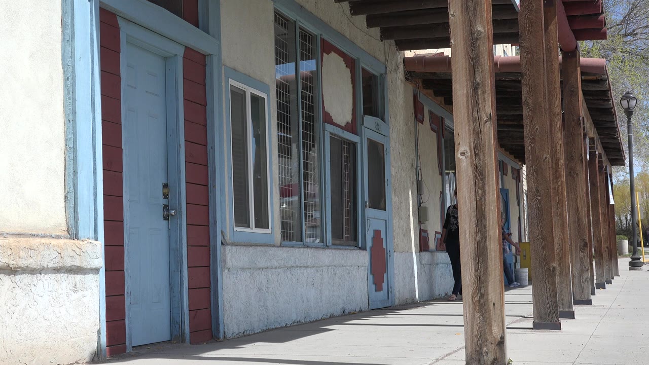 Opening view of a Taos, New Mexico apartment facade with a blue door at left, stucco wall, window trim, wooden porch posts, and no visible people.