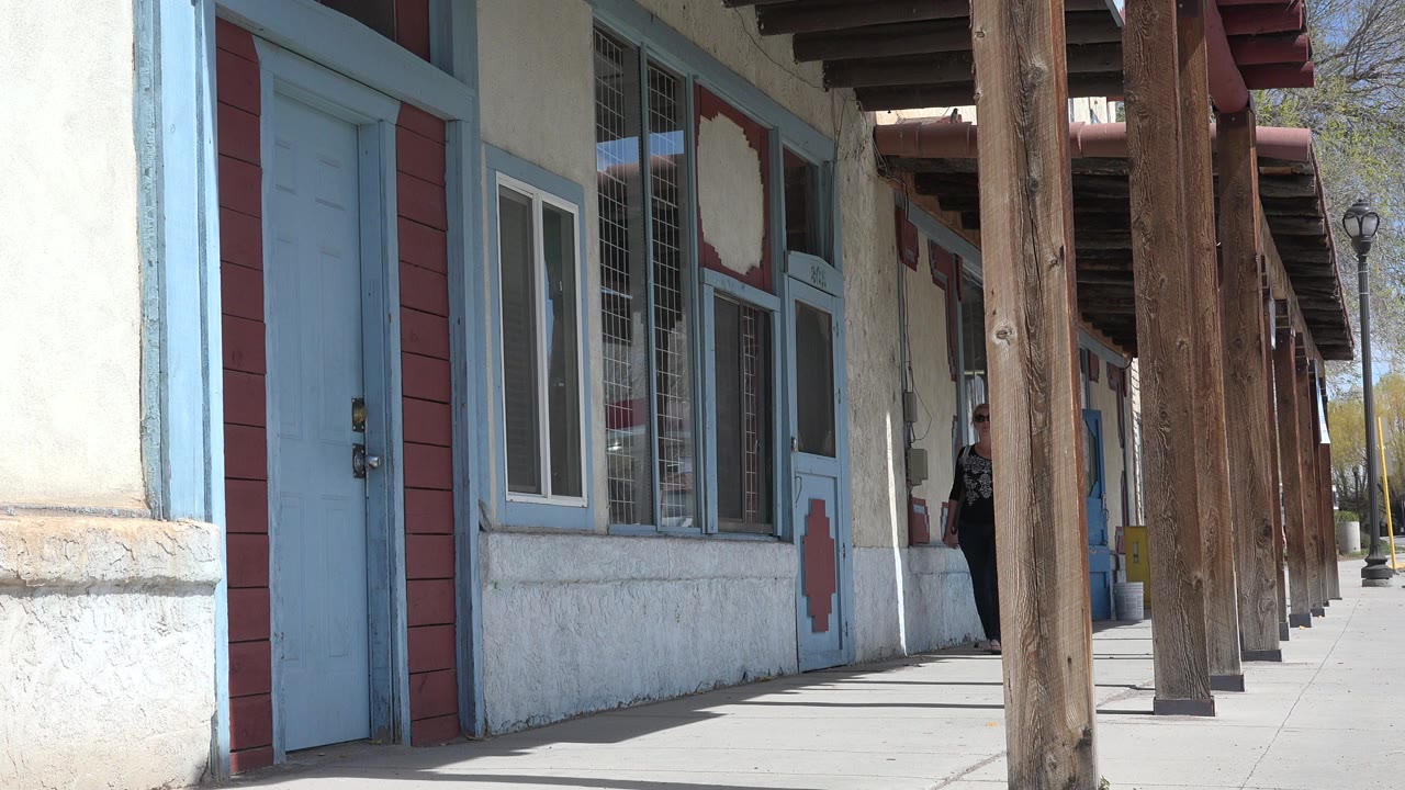 A woman in a black blouse and jeans steps into clearer view on a Taos, New Mexico sidewalk lined with blue doors, window grilles, and wooden posts.