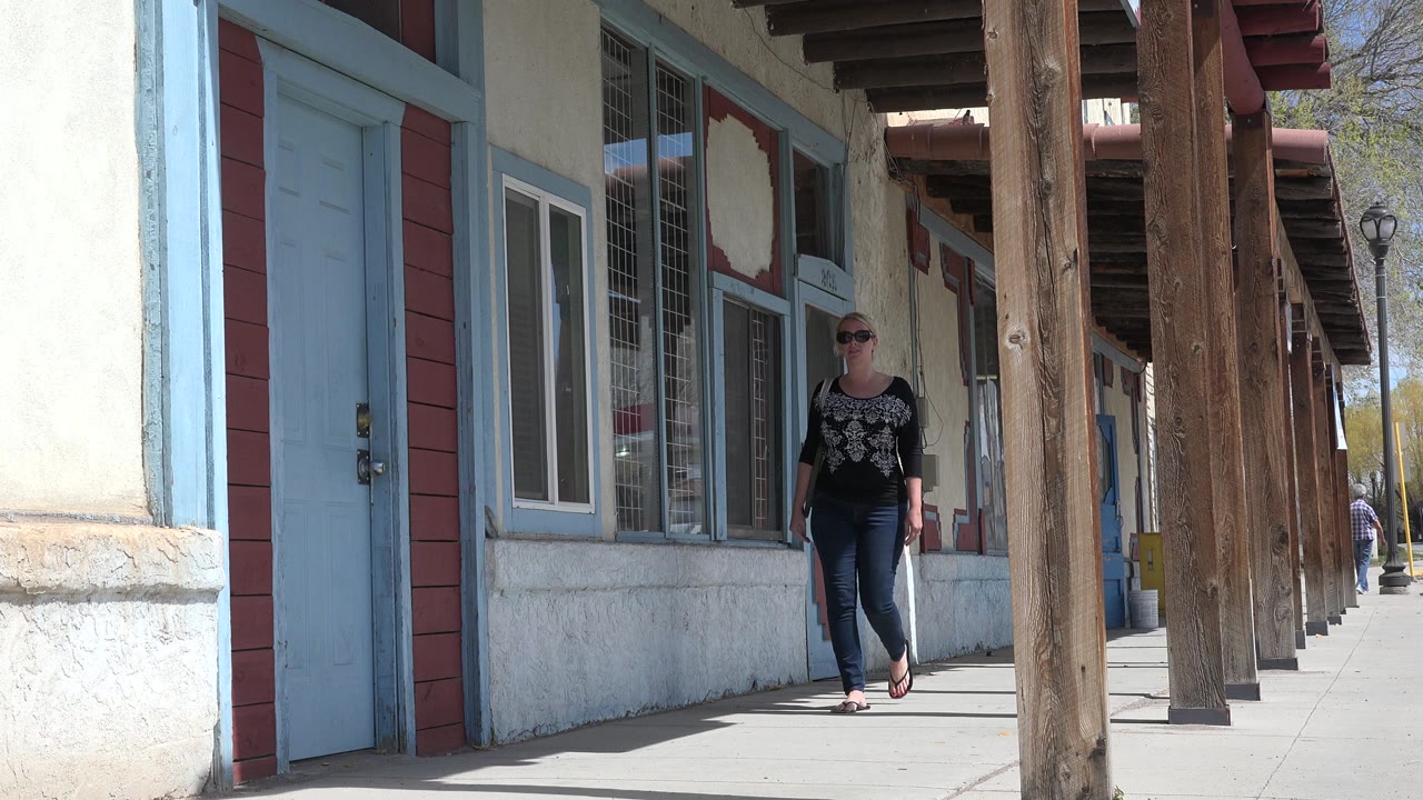 The woman advances along the Taos, New Mexico walkway, still mid-distance between the blue storefront door and the row of timber posts.