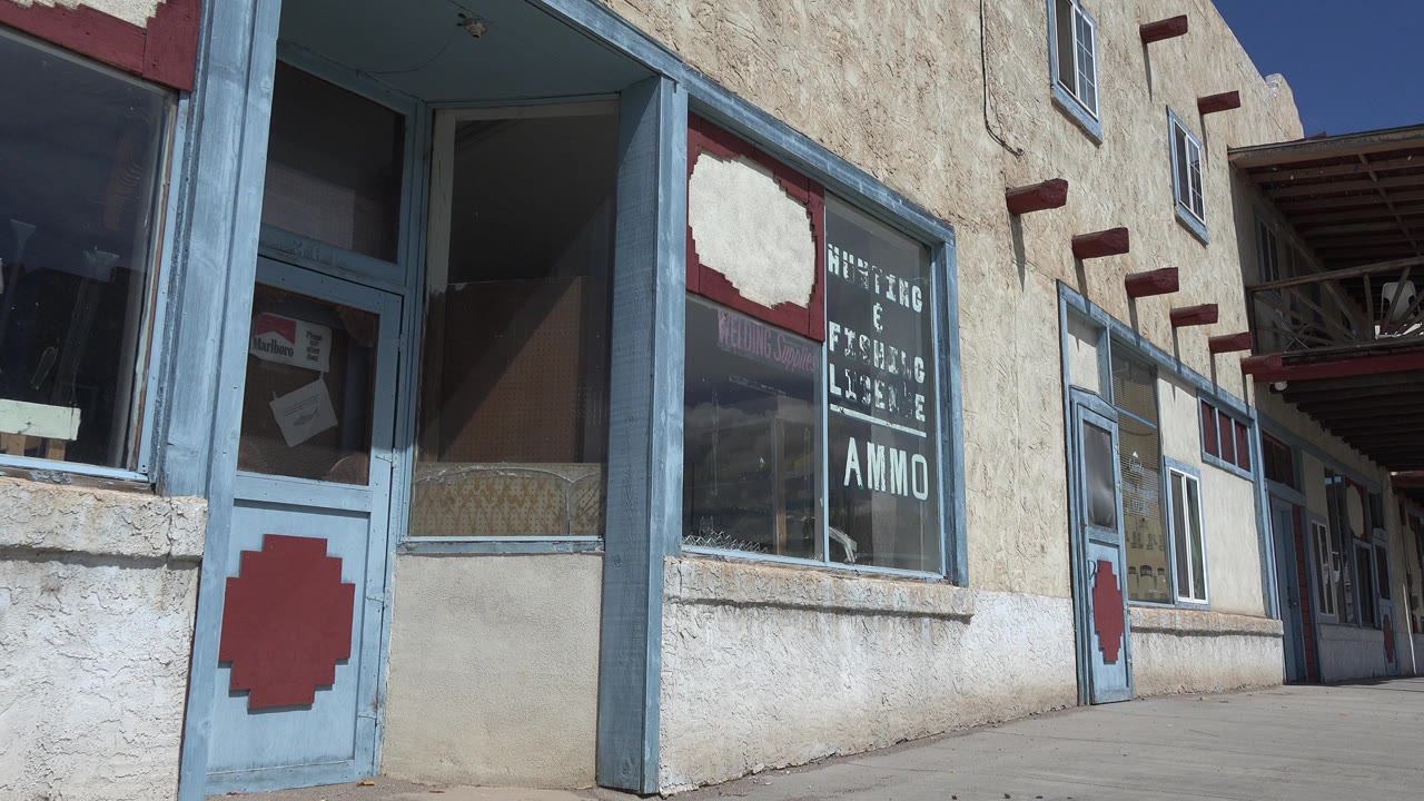 Opening frame of a blue-trimmed white stucco shop building in Taos, New Mexico, with glass doors, window lettering, red accents, bright sun, and no visible people.