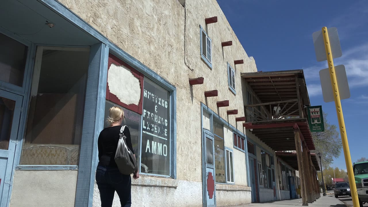 Woman seen from behind walking beside a storefront window on a sunny sidewalk.