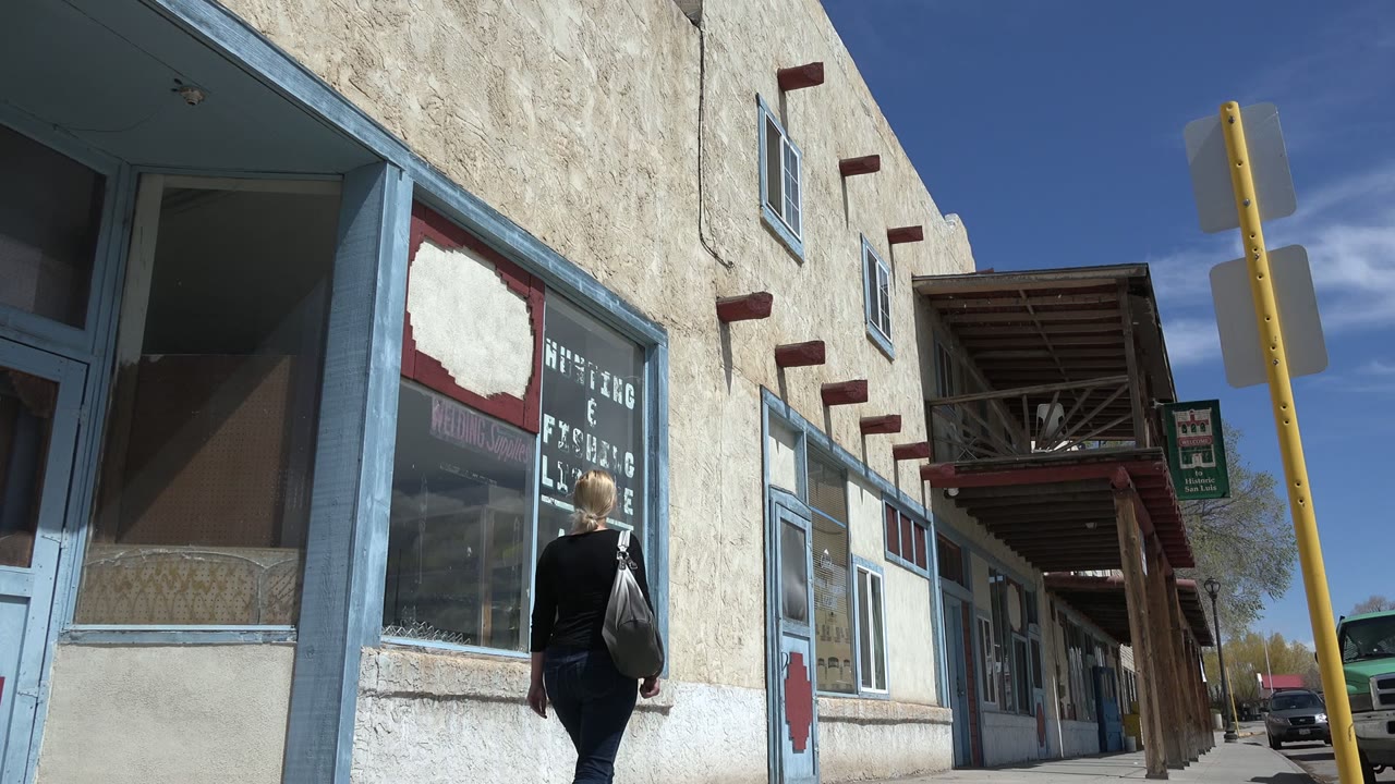 At 0:01 in Taos, New Mexico, the woman has moved farther along the storefront window where AMMO lettering is prominent and the covered walkway recedes ahead.