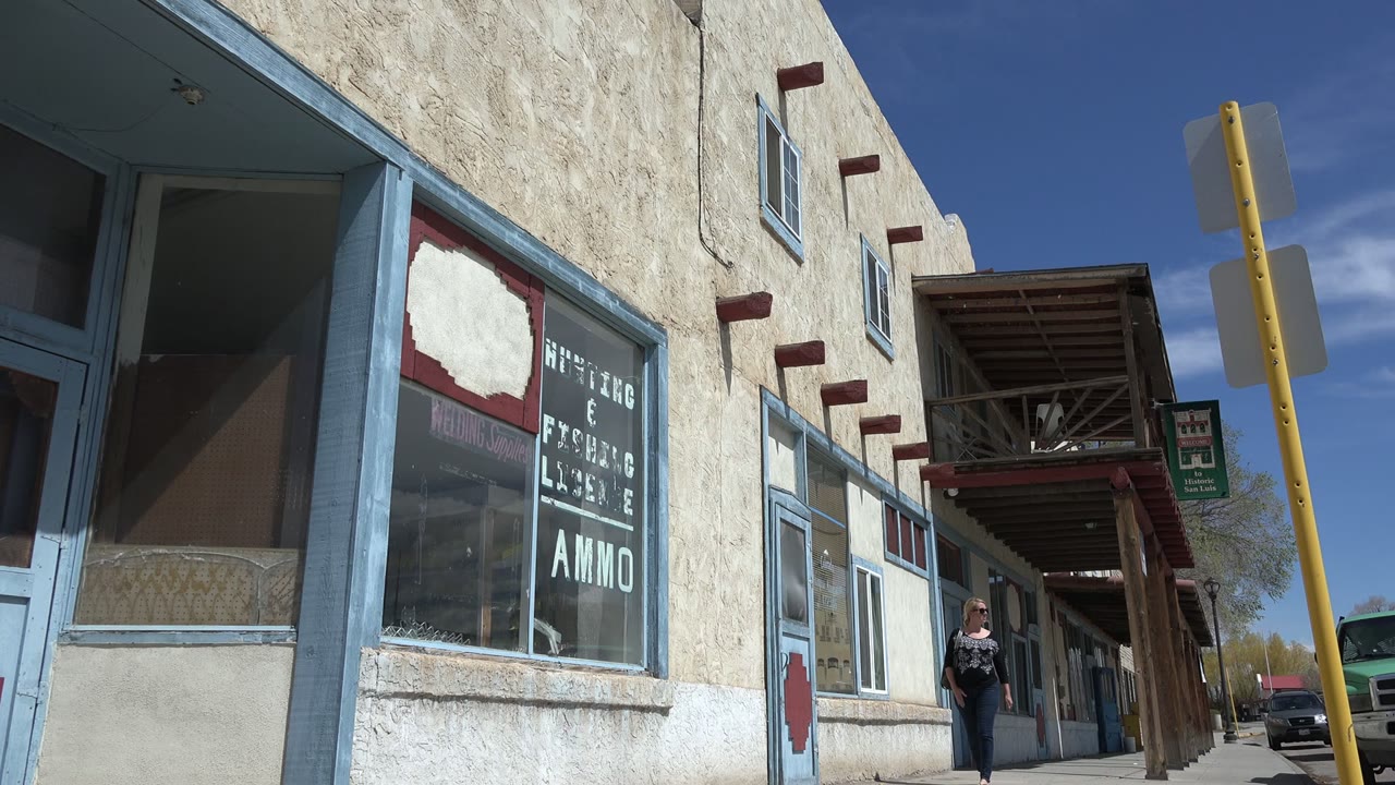 A woman in sunglasses walks at the edge of a Taos New Mexico storefront row, with blue-trimmed windows, stucco walls, and a shop window reading ammo visible in bright sun.