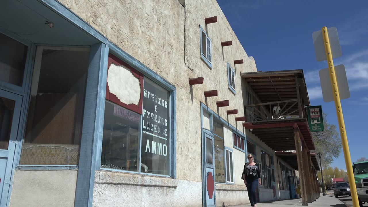 The woman has moved closer along the Taos New Mexico sidewalk, passing a large storefront window with white lettering while wood awning posts and parked vehicles line the background.