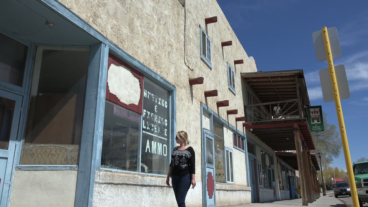 The woman moves closer to the center of the Taos New Mexico storefront view, passing window text that includes visible ammo lettering.