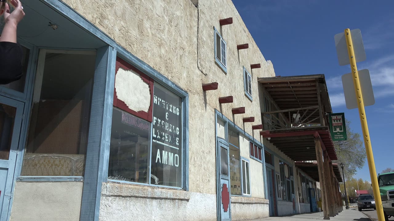The Taos New Mexico sidewalk view shifts away from the pedestrian to a stucco storefront with blue window trim, window lettering, a yellow post, and no visible people.