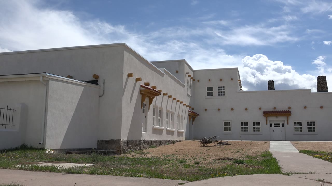 Opening frame of white cube-like buildings in Taos, New Mexico, with a sidewalk, dirt foreground, grass edge, bright clouds, and no visible people.