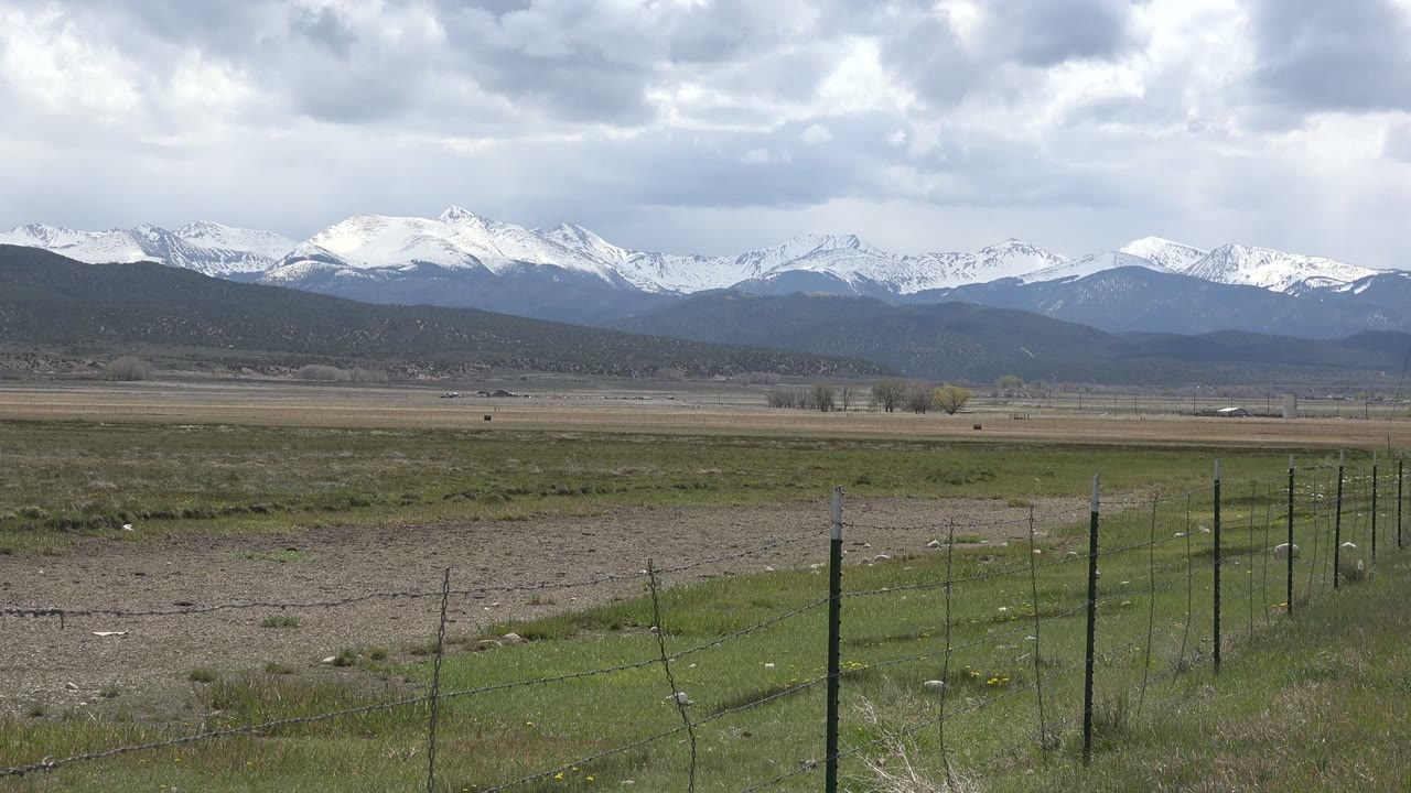 Wide grassy field with fence posts, distant snowy mountains, and cloudy sky.