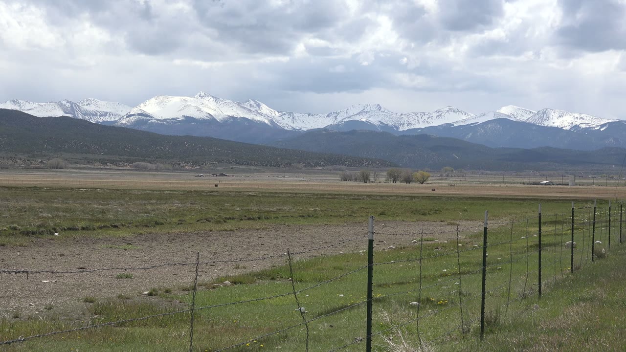 Snowy mountains beyond grassland and a wire fence under cloudy sky.