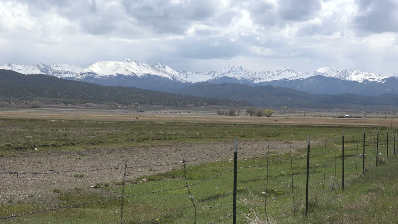 Open field with fence posts, distant snowy mountains, and cloudy sky.
