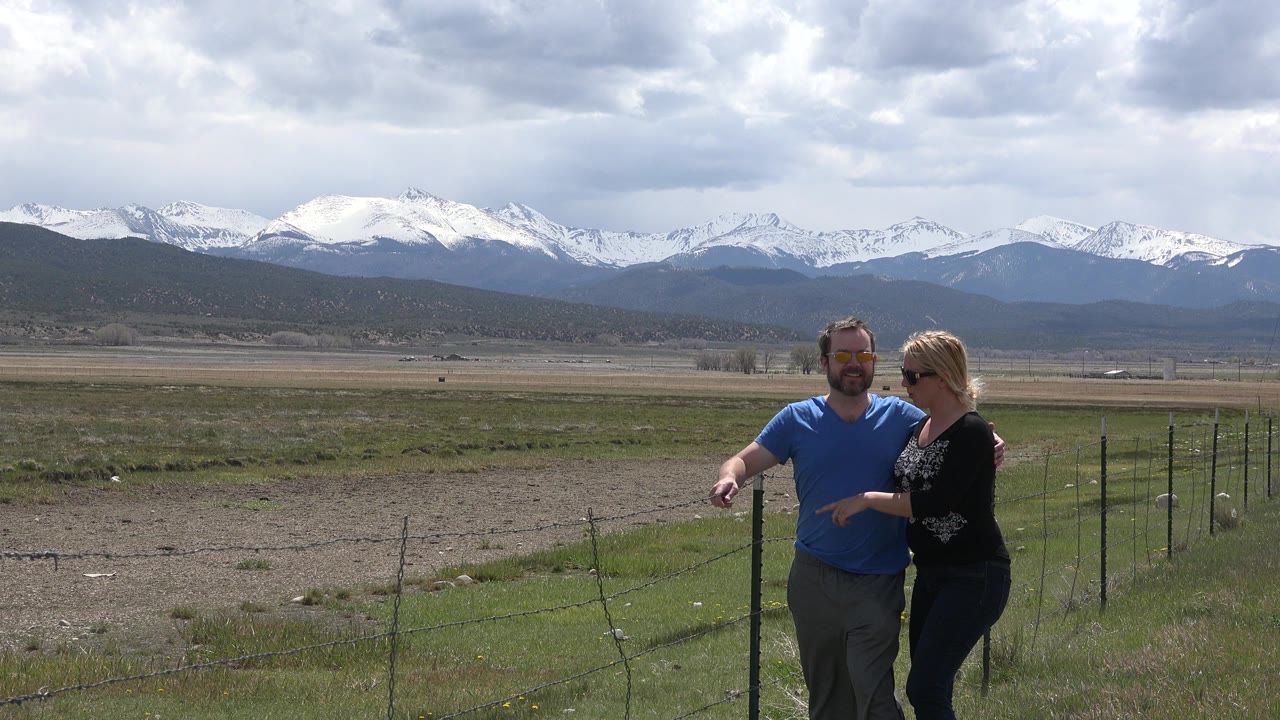 At 0:01, the couple remains close together beside the fence line in Taos New Mexico, with the man's arm angled forward and the woman's sunglasses visible against the bright valley backdrop.