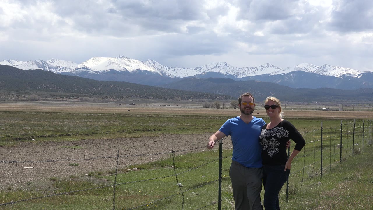 Man and woman standing by a fence with snow-covered mountains behind them.