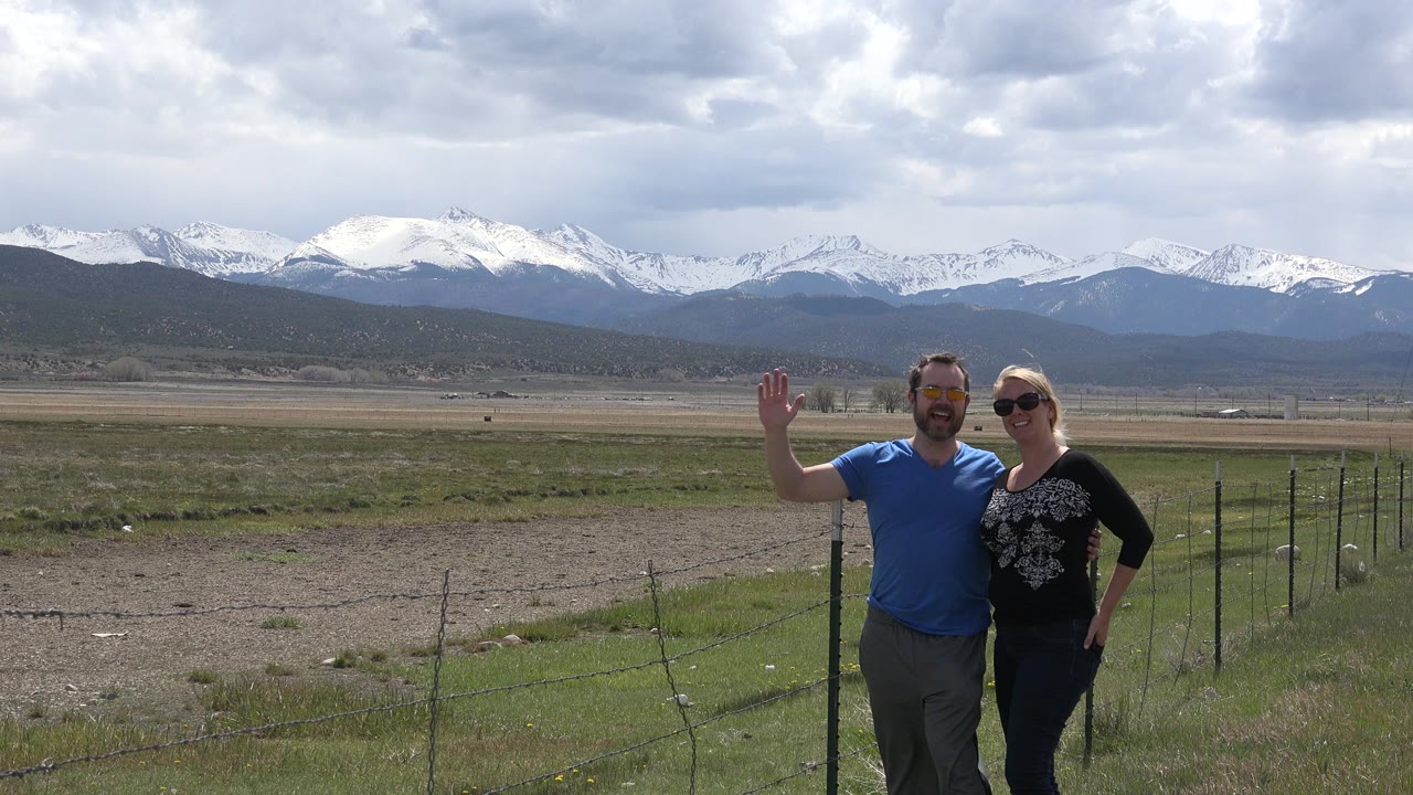 Two adults posing near a fence with snow-capped mountains behind them.