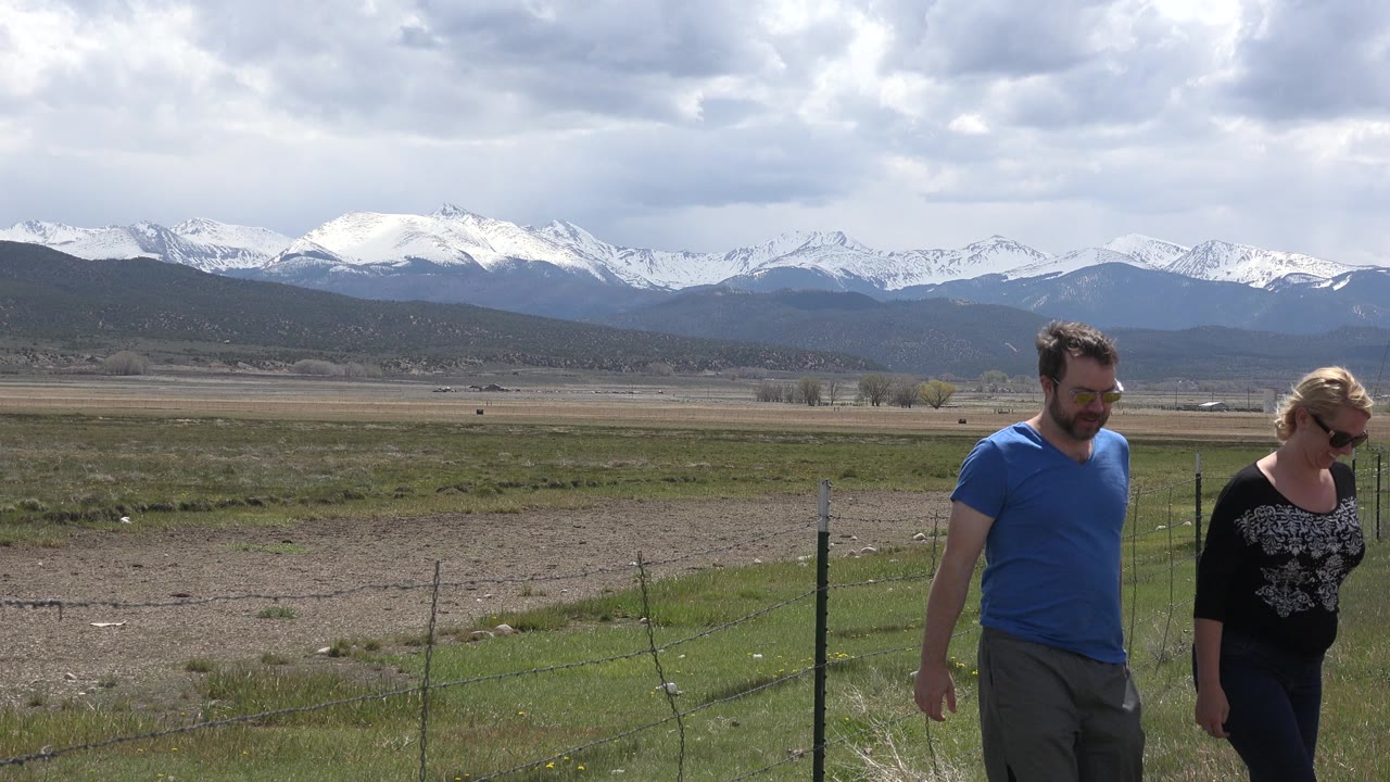 Two adults walking beside a wire fence with snowy mountains in the distance.