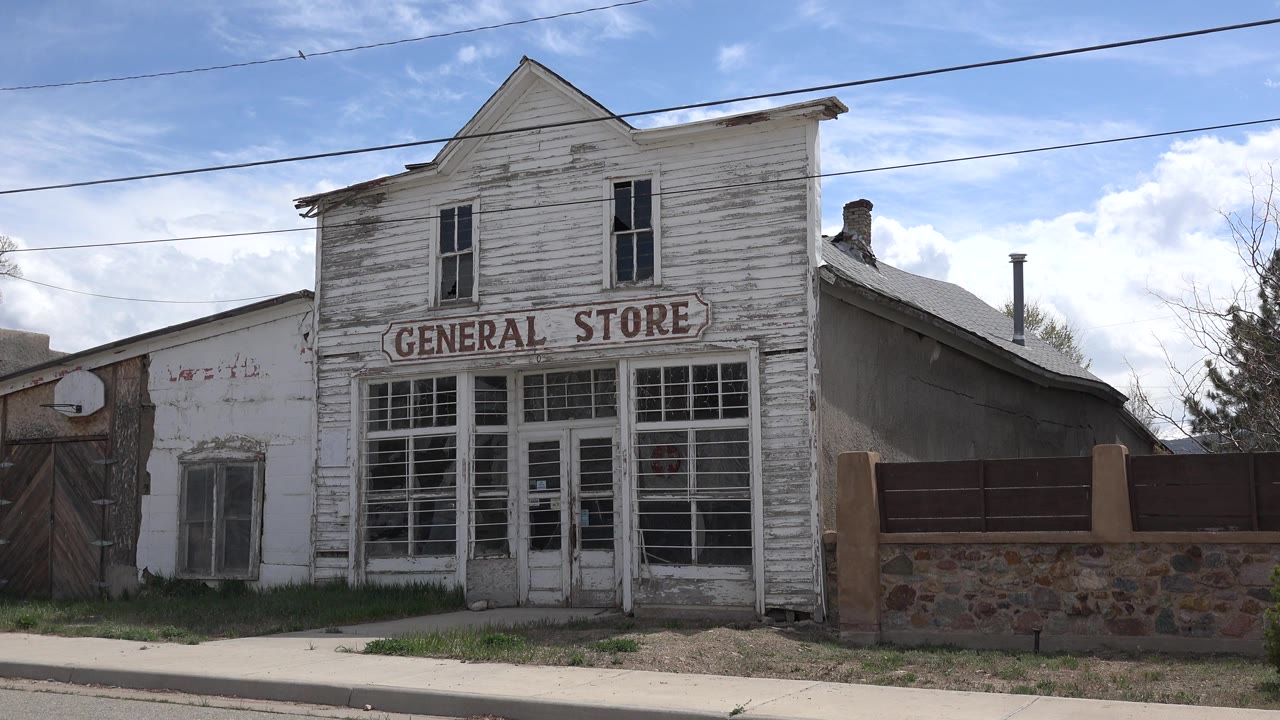 Opening frame of a weathered white wooden general store in Taos, New Mexico, with red lettering above gridded windows, overhead wires, daylight, and no visible people.