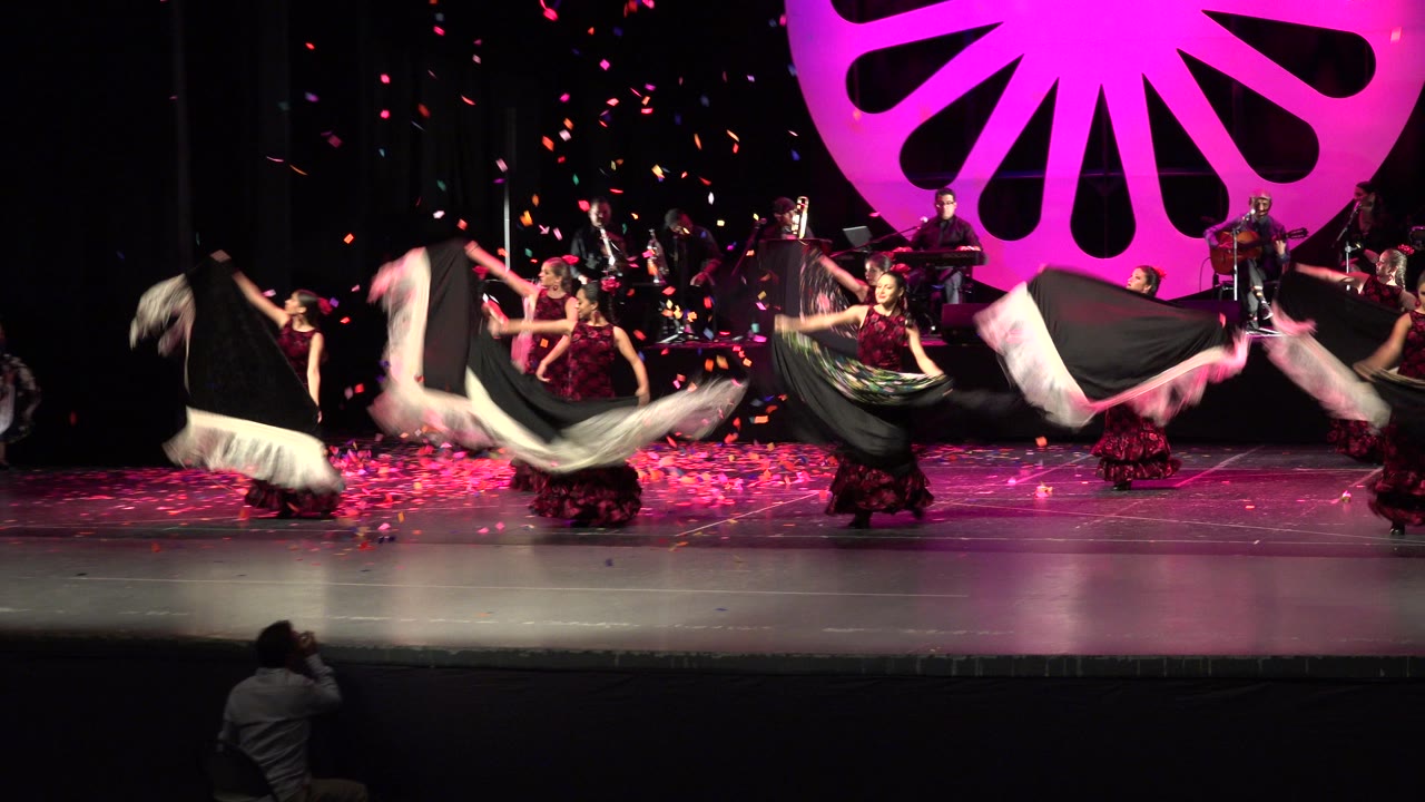 Women perform a choreographed dance on a Cabo San Lucas stage, lifting white and dark fabric panels while confetti falls near a large pink backdrop.