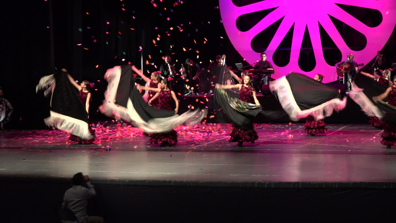 The Cabo San Lucas performance continues with women in dark red dresses sweeping white and black cloth across the stage in front of musicians and a bright pink set piece.