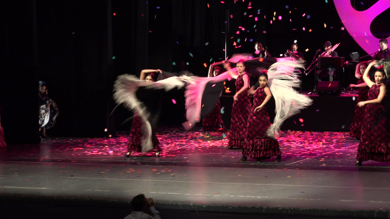 Stage dancers in burgundy dresses perform as petals fall near a live band.