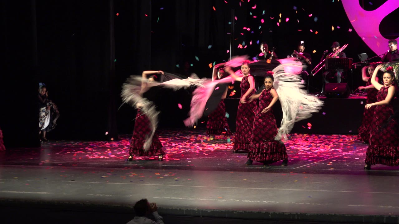 In Cabo San Lucas, Mexico, several dancers hold poses while another sweeps white fringed fabric across the stage under falling colored petals.