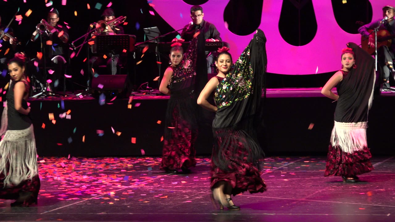 Female dancers in black and red costumes perform on a pink-lit stage in Cabo San Lucas, Mexico, with confetti falling and musicians seated in the background.