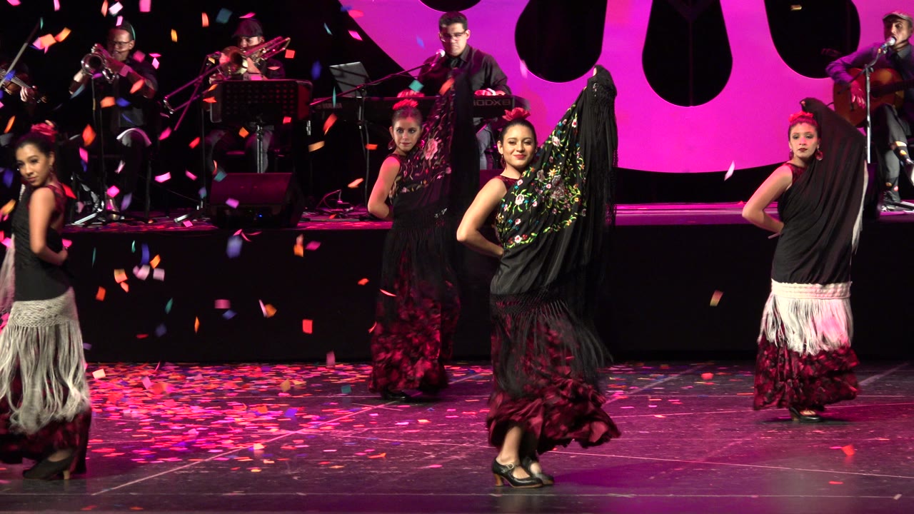Three female dancers hold positions across the stage in Cabo San Lucas, Mexico, while pink and orange confetti hangs in front of the musician platform.