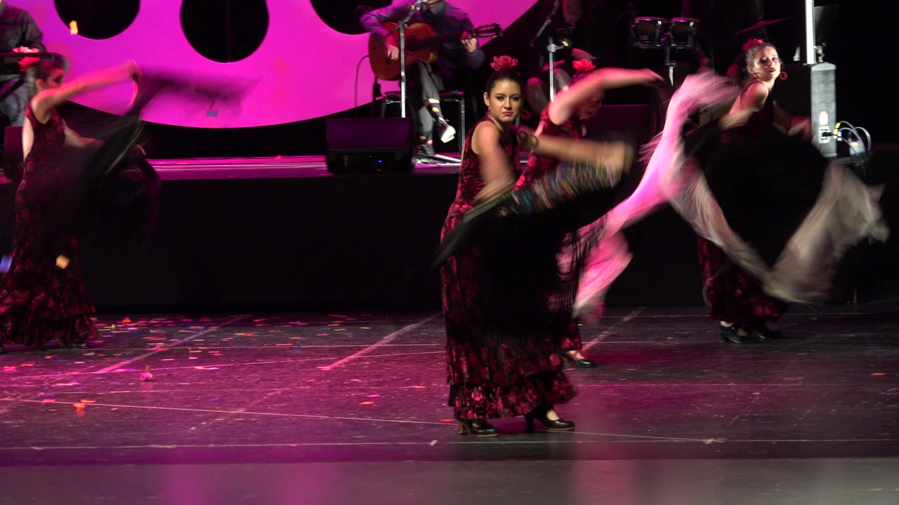 In Cabo San Lucas, Mexico, dancers in red and black stage dresses perform with fringed shawls while magenta lighting fills the backdrop and musicians sit behind them.