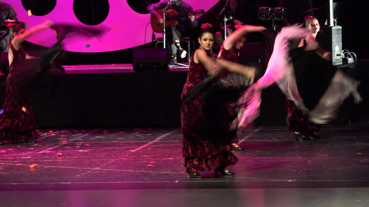 In Cabo San Lucas, Mexico, a central dancer faces the audience as other performers blur through shawl movements against a bright pink stage background.