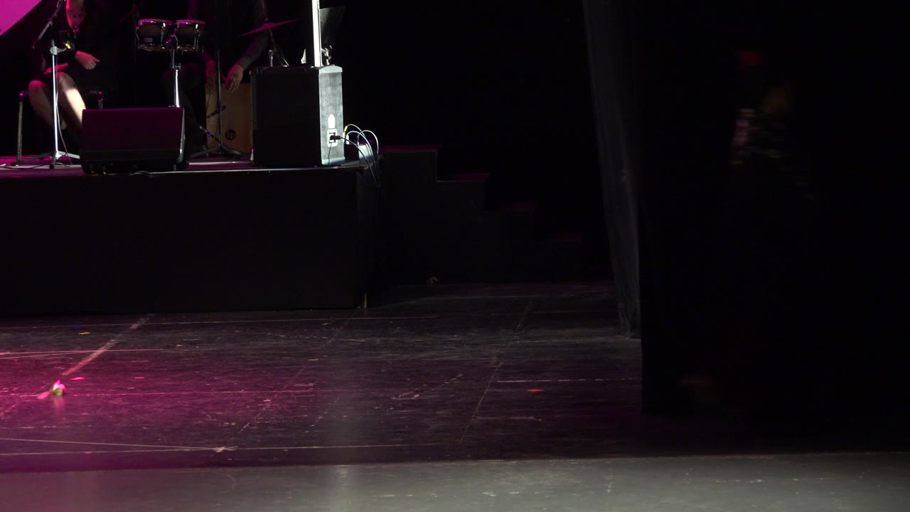 The Cabo San Lucas performance stage remains distant, with pink light at the top, equipment on the platform, and a wide dark floor dominating the frame.