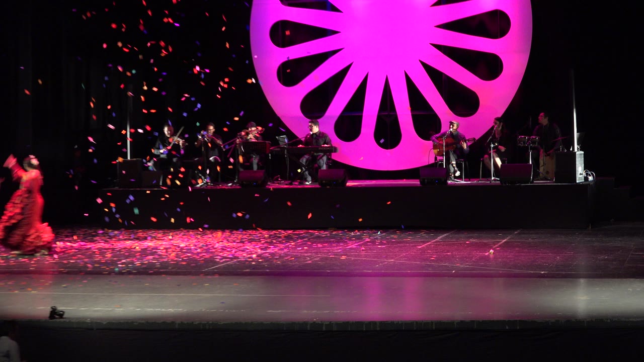 Wide stage with orange-dressed dancer, band, and pink circular backdrop.
