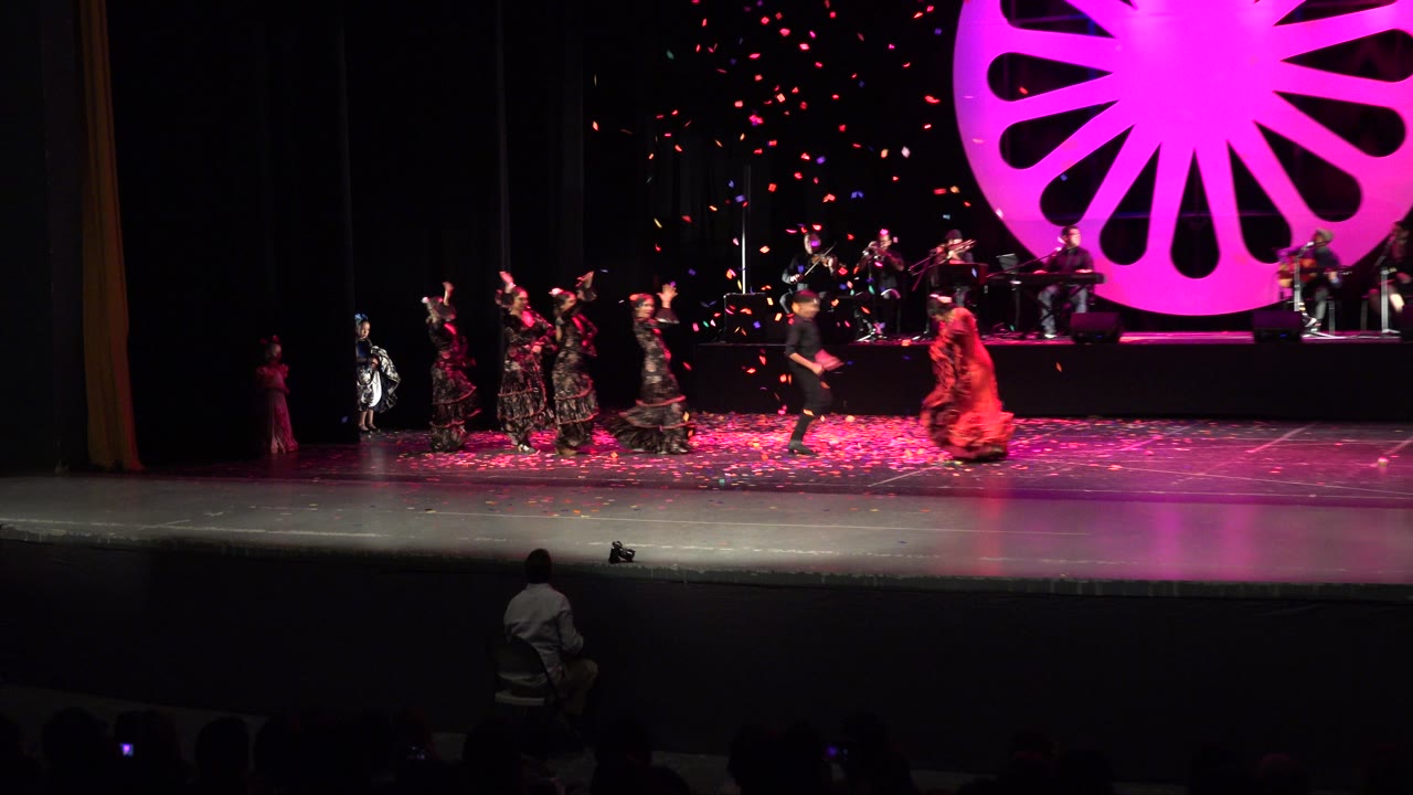 Wide stage view with dancers, musicians, pink backdrop, and confetti.
