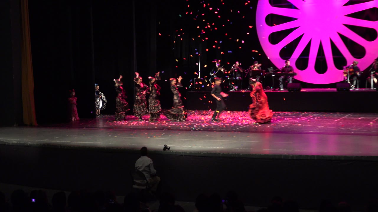 Cabo San Lucas performers continue across the stage as a red-dressed dancer moves near center, with musicians behind and pink light filling the backdrop.