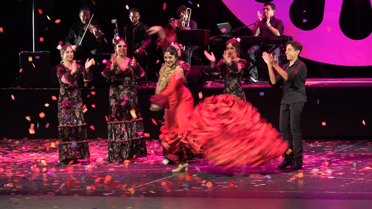 Red-dressed dancer spins on a stage as performers clap around her.