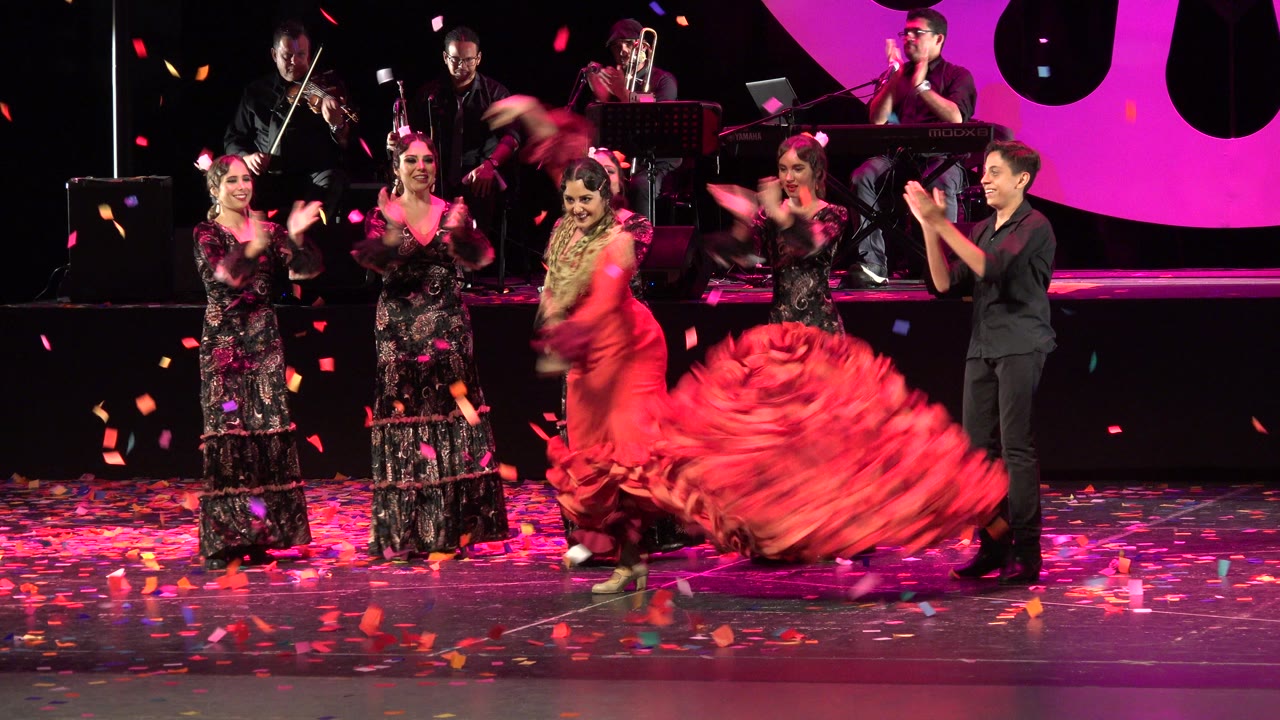 In Cabo San Lucas, Mexico, the red-dressed dancer sweeps her skirt low across the stage as clapping performers line the midground and musicians remain visible on the rear platform.