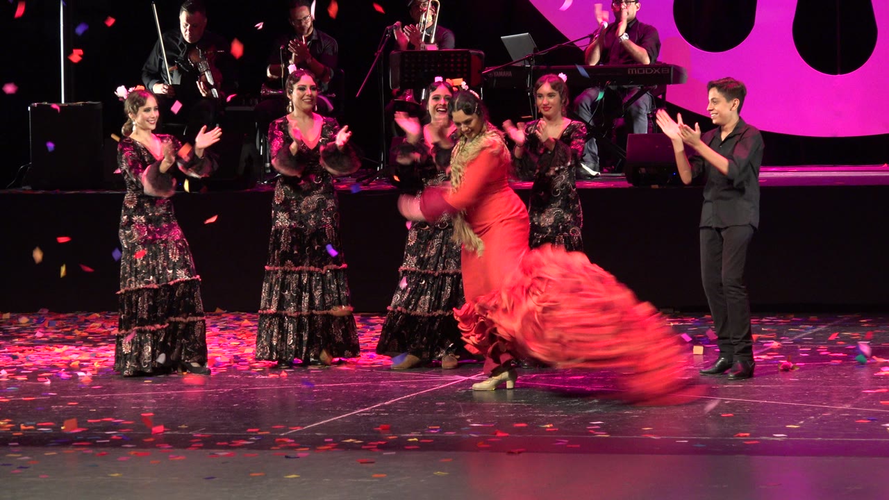 Dancer in a red ruffled dress spins on a stage with performers clapping nearby.