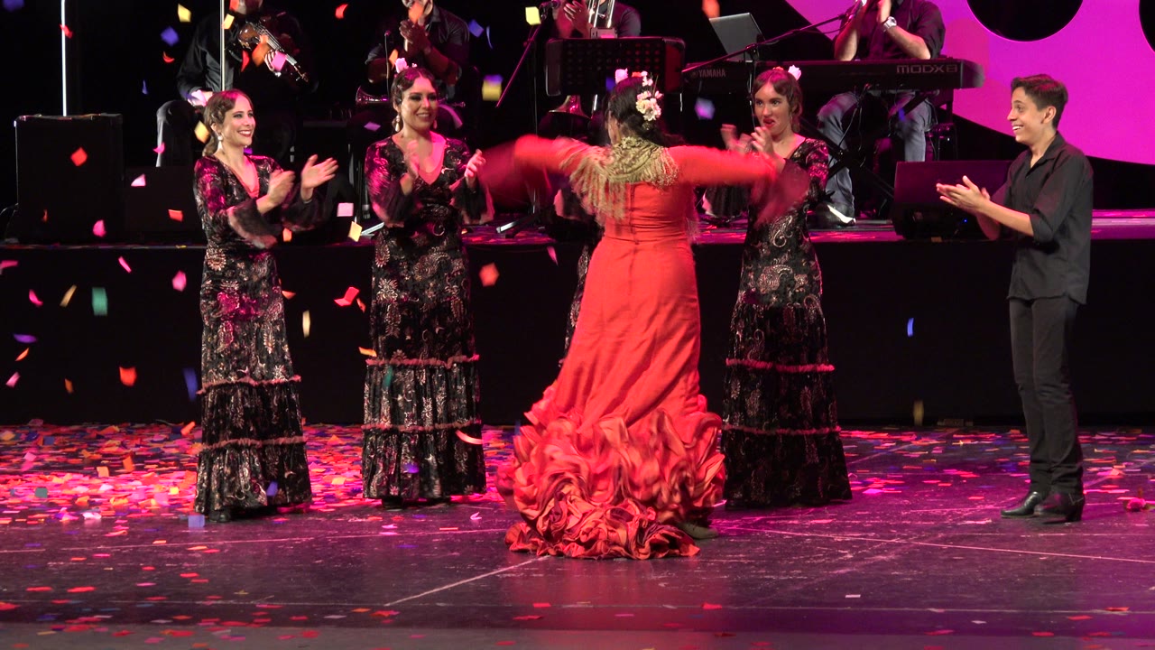 Stage dancers clap around a performer in a red ruffled dress.