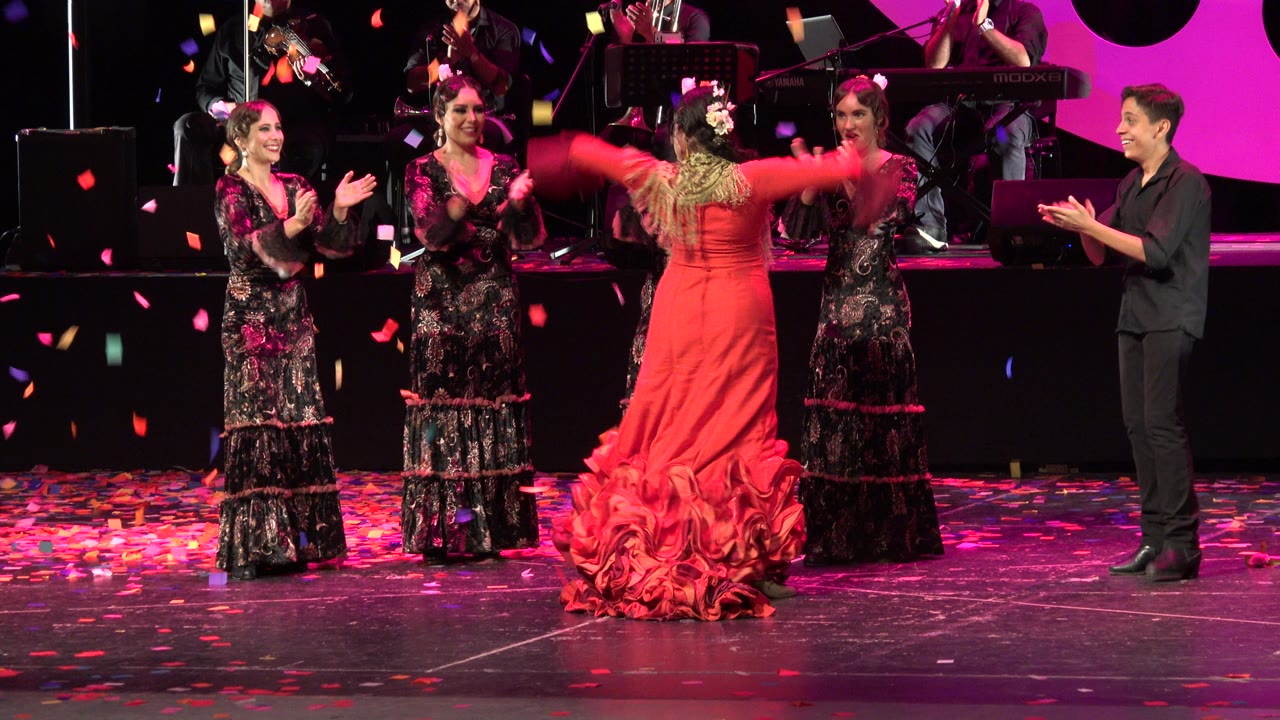 In Cabo San Lucas, Mexico, the red-dressed dancer faces the clapping performers while confetti lies across the stage and the band remains visible in the background.