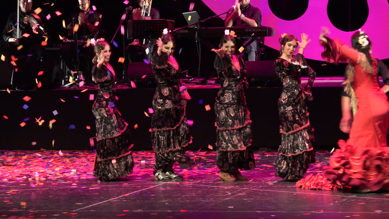 A Cabo San Lucas Mexico 2019 stage performance frame shows women in black patterned dresses dancing as confetti falls, with musicians behind them and a red-dressed performer blurred at right.