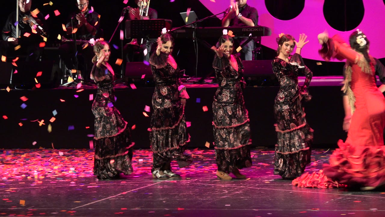 Women in black and red stage costumes perform under magenta lighting in Cabo San Lucas Mexico, with confetti suspended in the air and a red ruffled dress crossing the right side of frame.