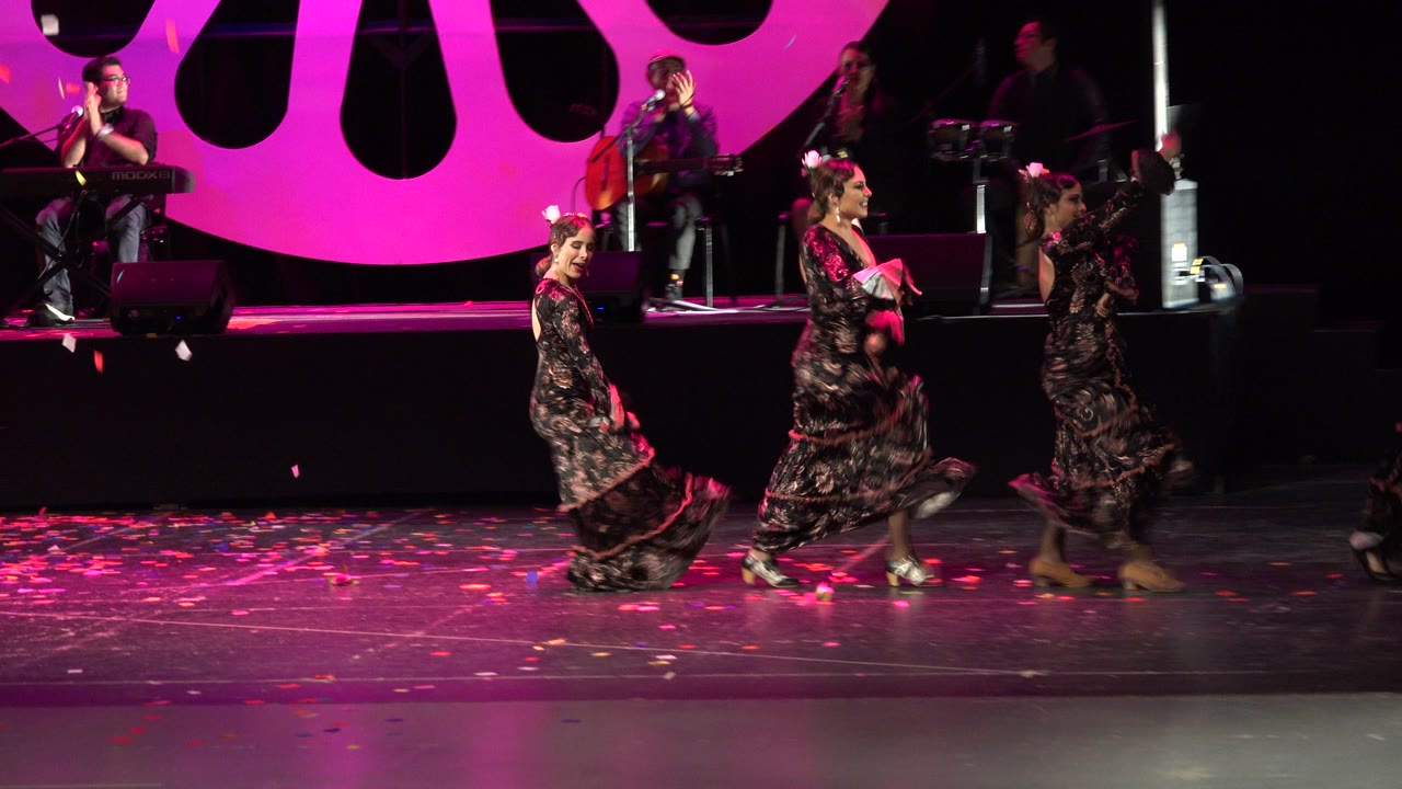 Three women dance on a magenta-lit stage with a live band behind them.