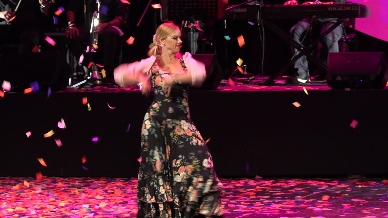 In Cabo San Lucas, Mexico, the woman turns slightly left in her floral dress while colored petals fall across the dark stage background.
