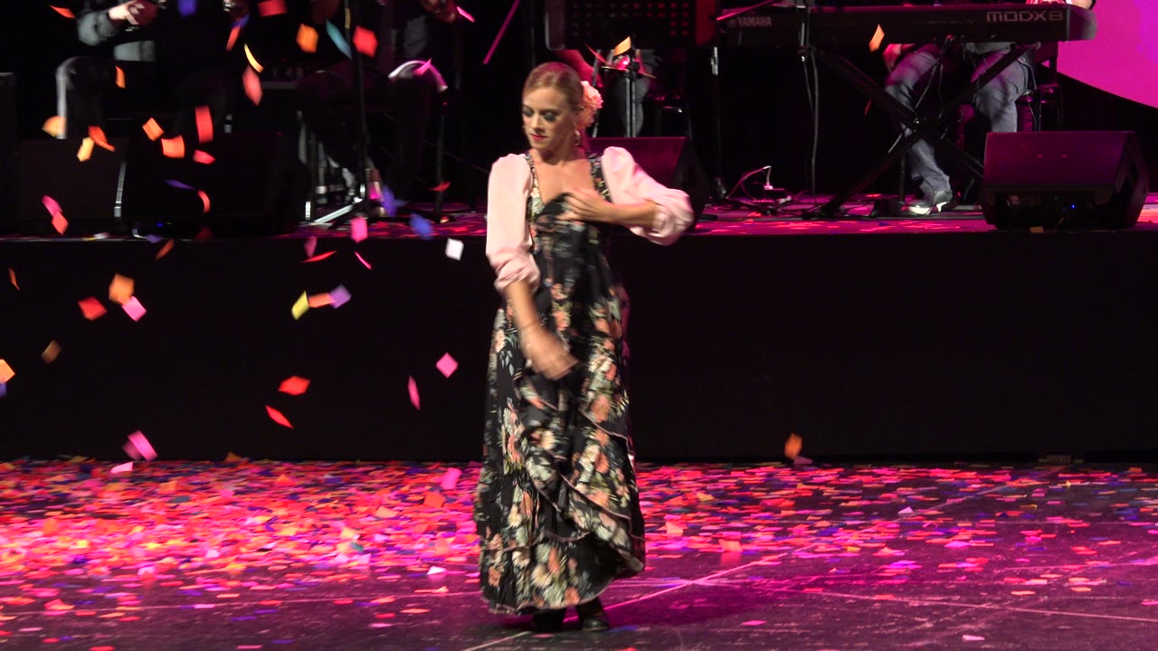 The dancer moves across the Cabo San Lucas stage with one arm lowered, colored confetti suspended in front of the band area and dark stage backdrop.